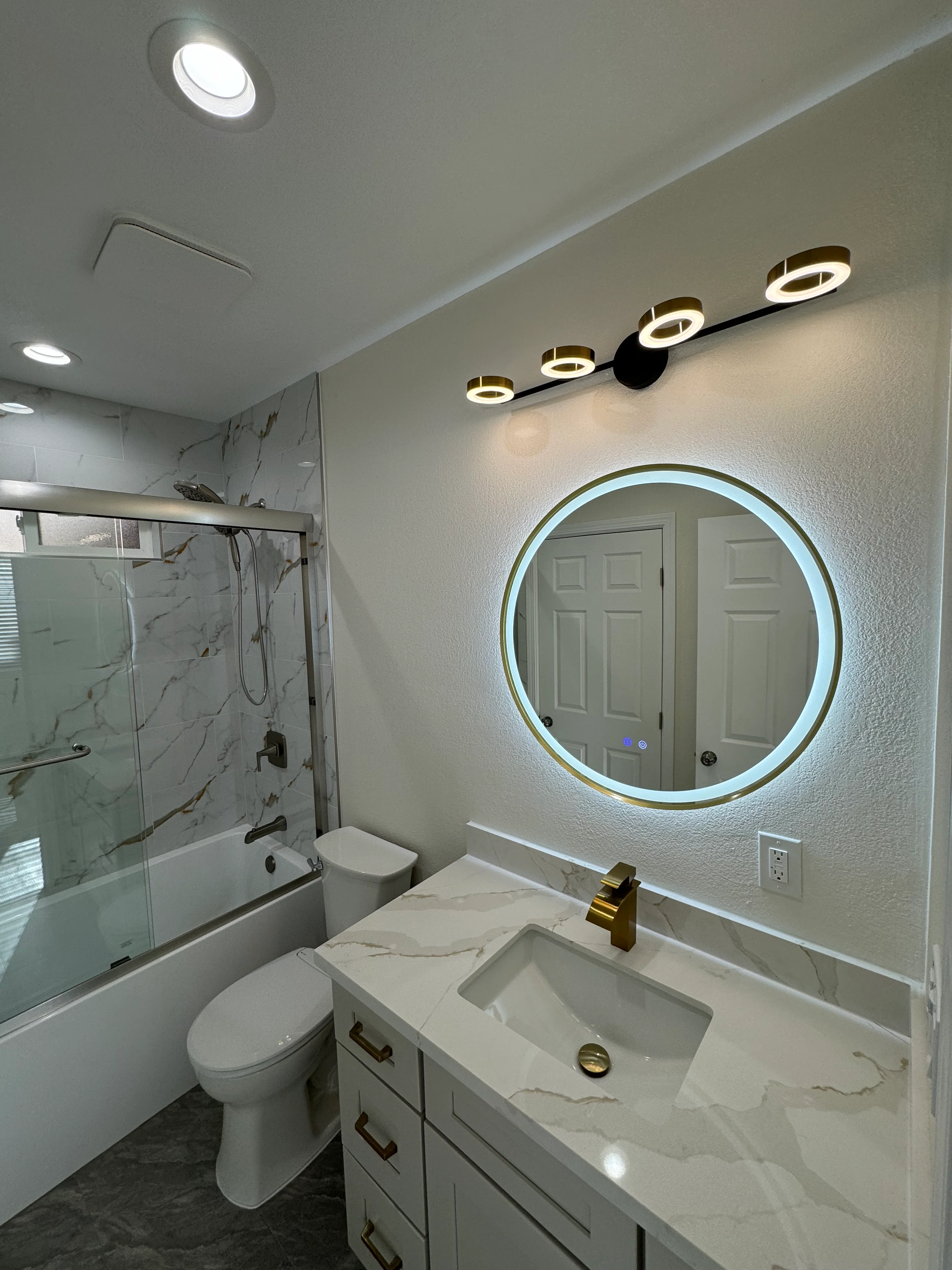 Modern bathroom with marble vanity, backlit mirror, brass fixtures, and glass shower enclosure