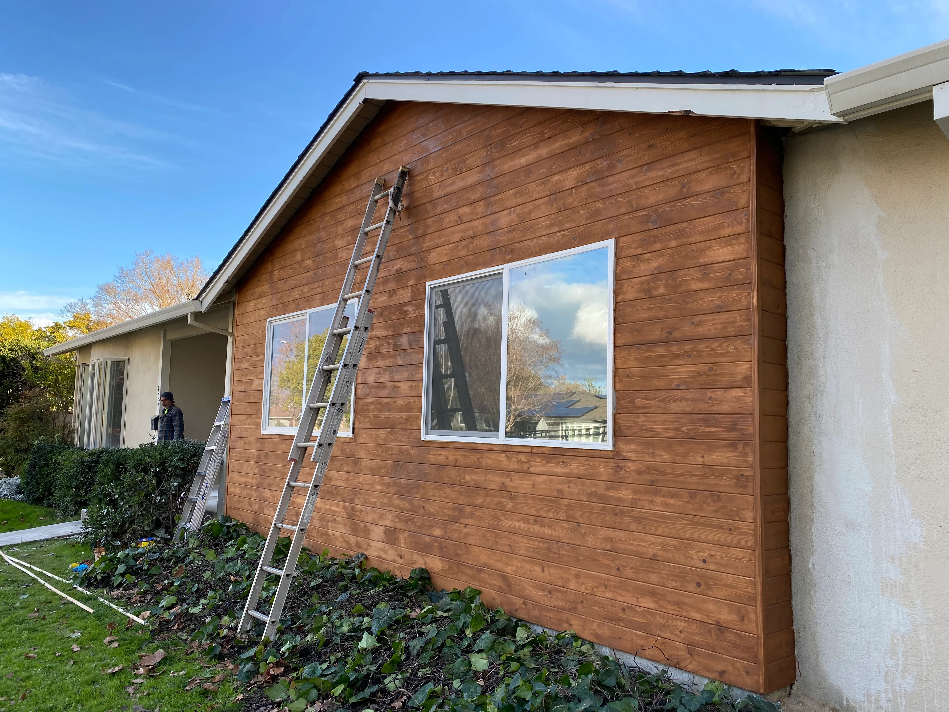 Brown horizontal wood siding with white-trimmed casement windows and metal roof installation