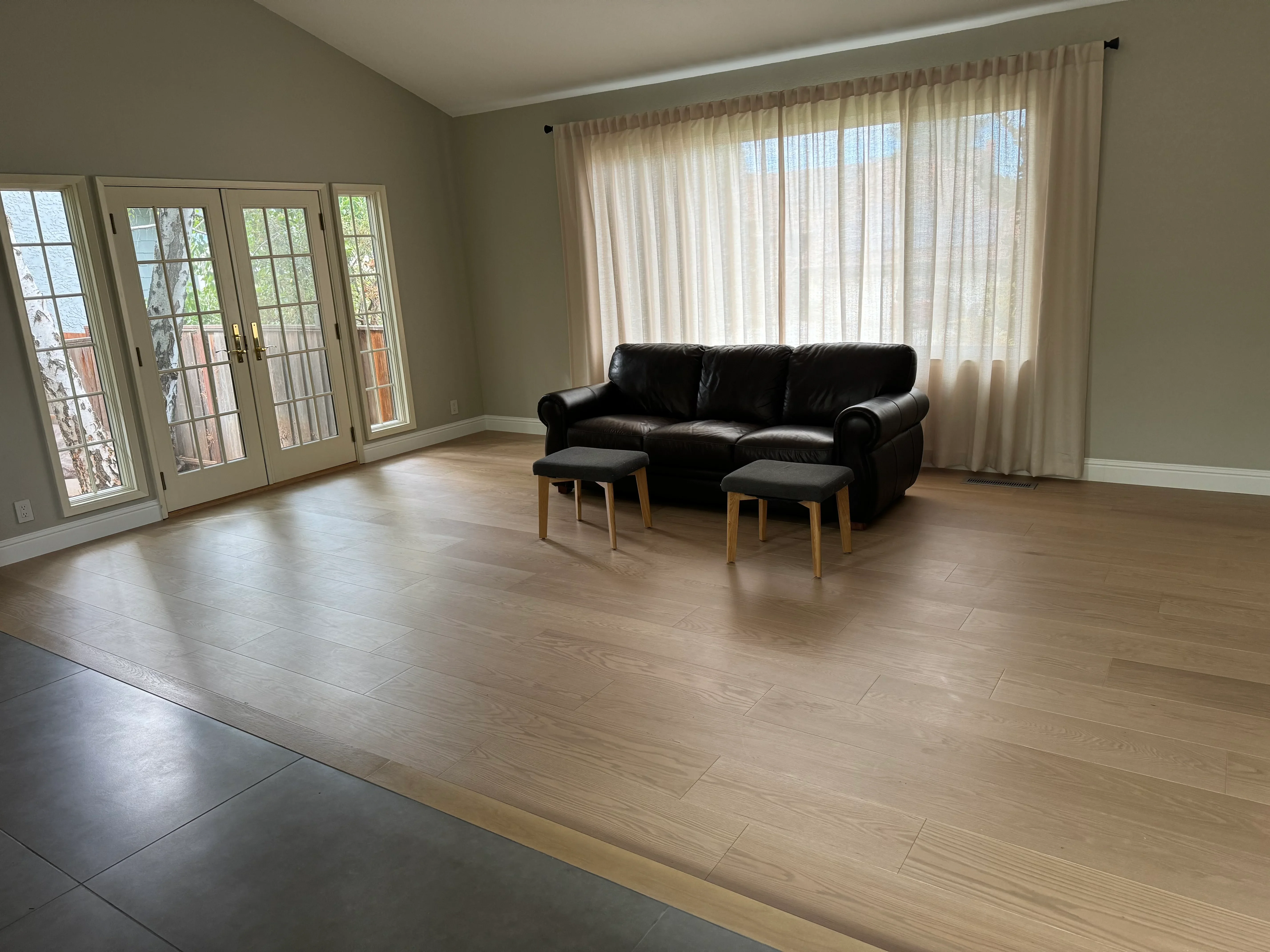 Bright living room with wood flooring, black leather sofa, French doors, and floor-to-ceiling windows with sheer curtains