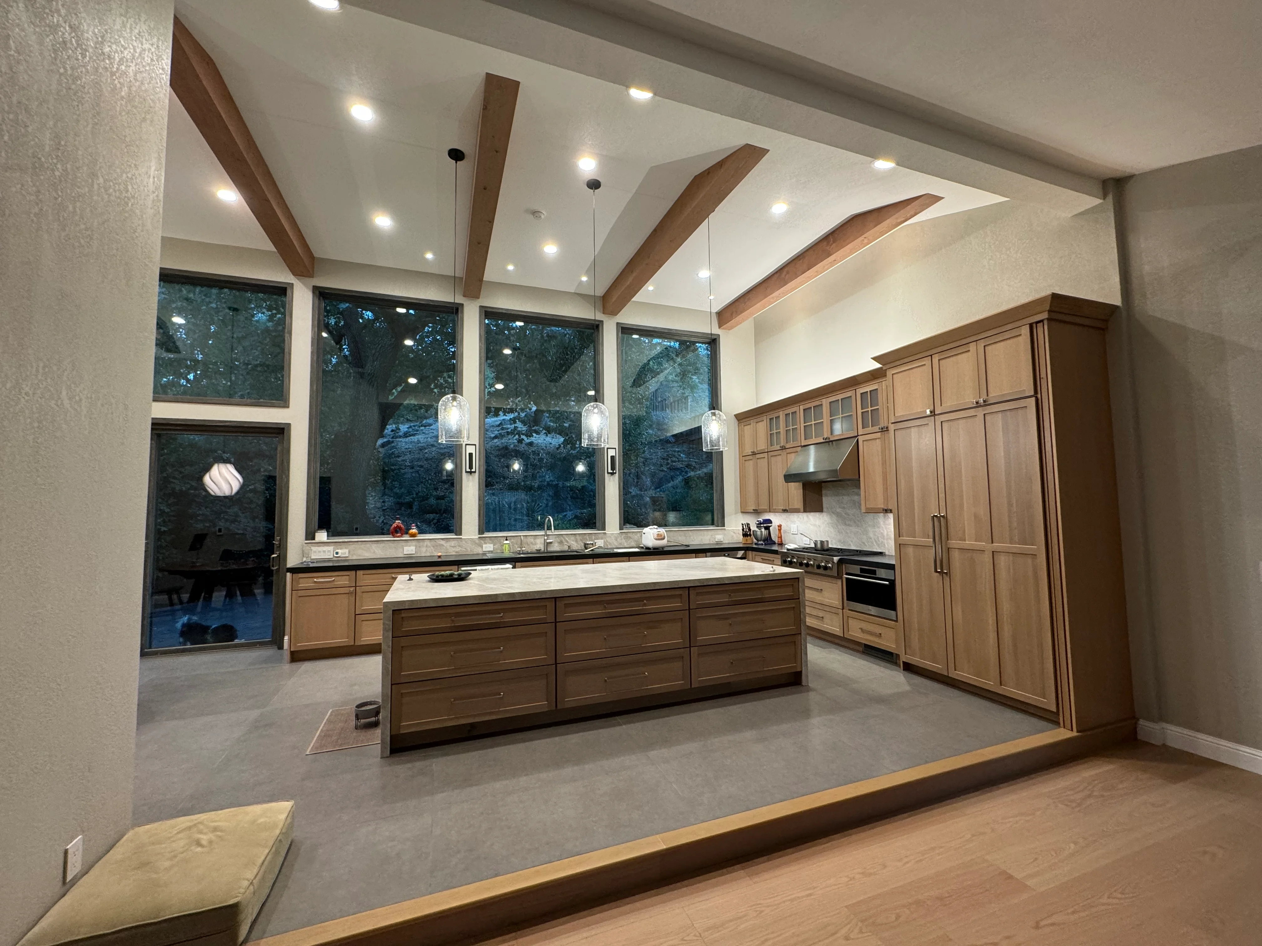 Modern kitchen with wood cabinetry, quartz counters, island, vaulted ceiling with beams, and large windows