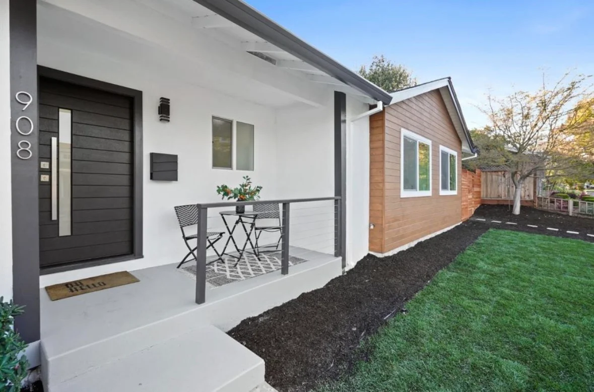 Modern home exterior with white stucco, dark siding, concrete patio, metal pergola, and landscaping
