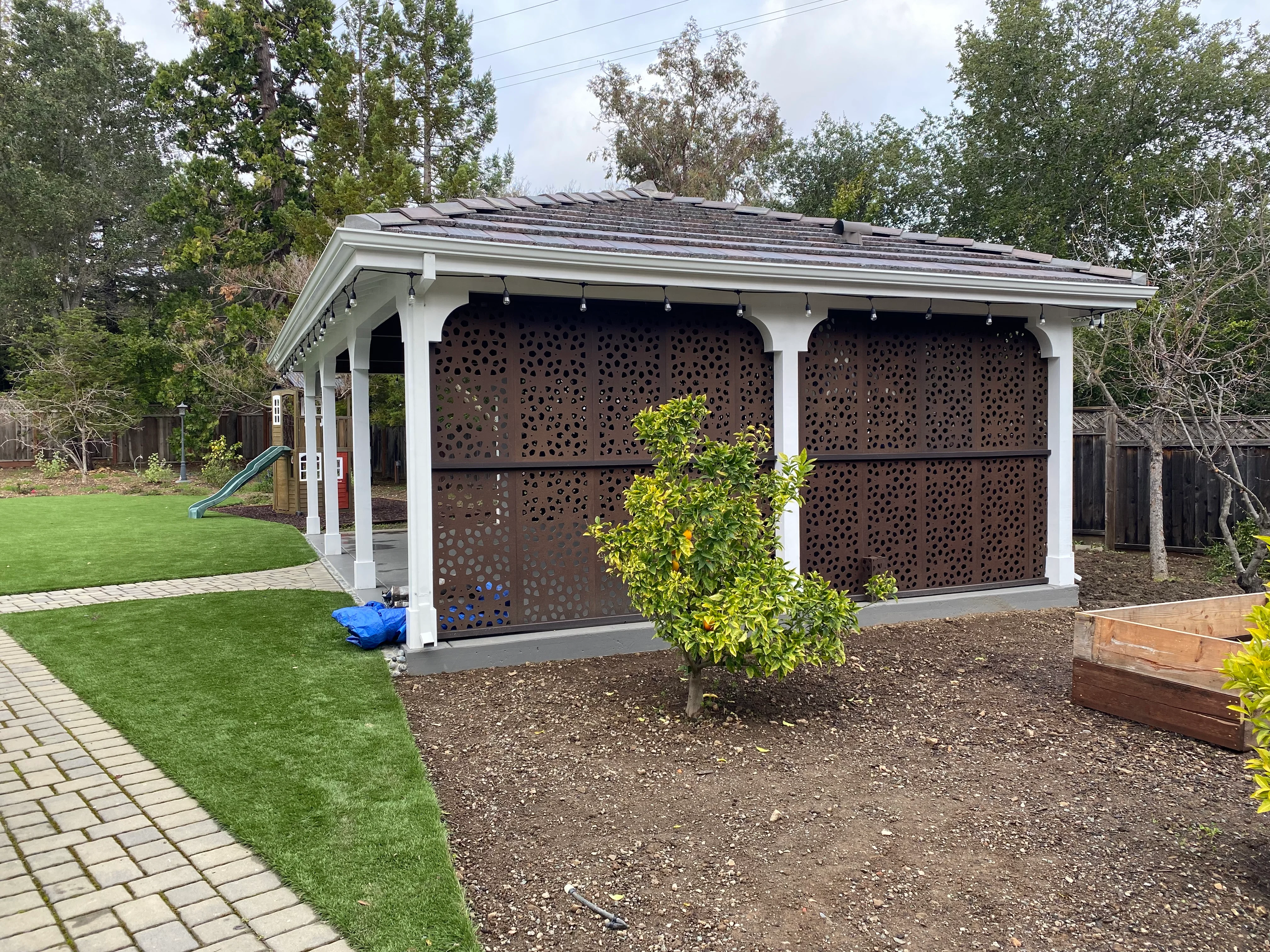 White pergola pavilion with bronze perforated metal screens, shingled roof, and pendant lighting in backyard setting