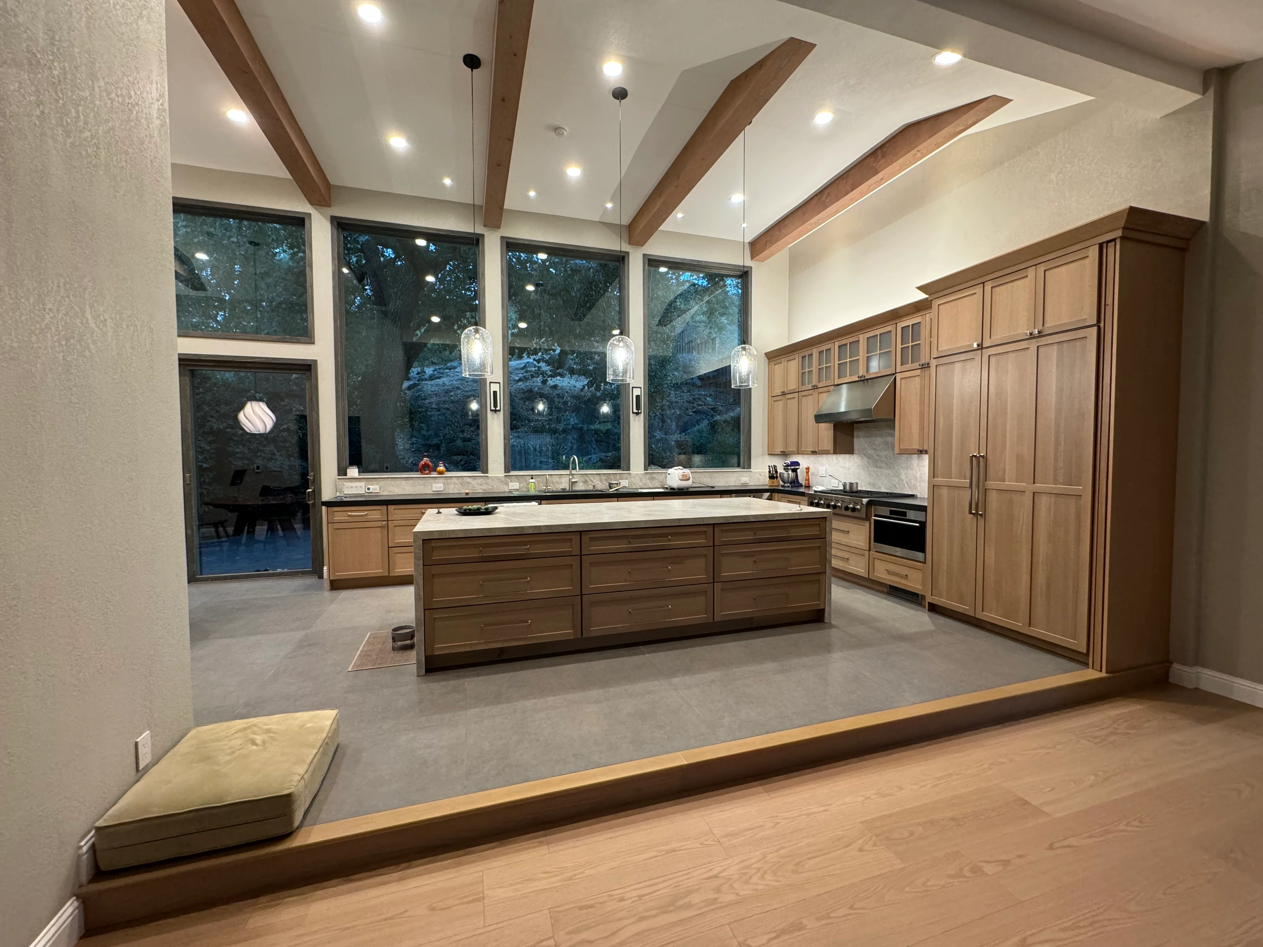 Modern kitchen with wood cabinetry, quartz counters, island, vaulted ceiling with beams, pendant lights, and large windows