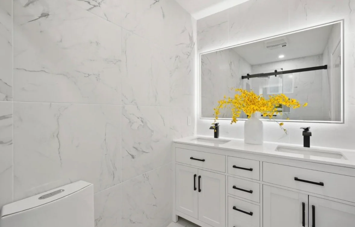 Modern bathroom with marble tile walls, dual sink vanity, frameless glass shower, and LED-backlit mirror