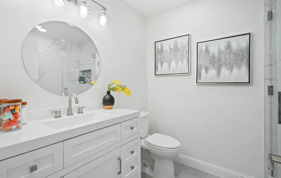 White bathroom vanity with quartz countertop, frameless glass shower, and circular mirror